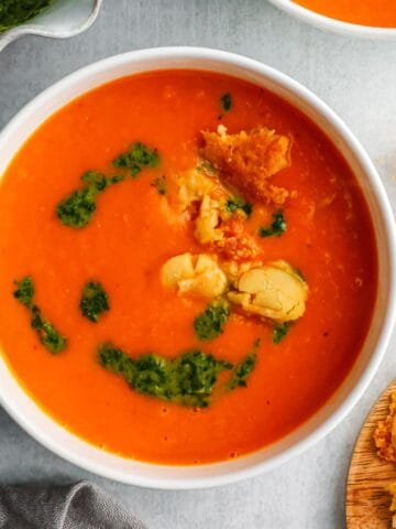 A white bowl filled with vibrant orange roasted pepper and white bean soup topped with green pesto and crispy parmesan crisps.