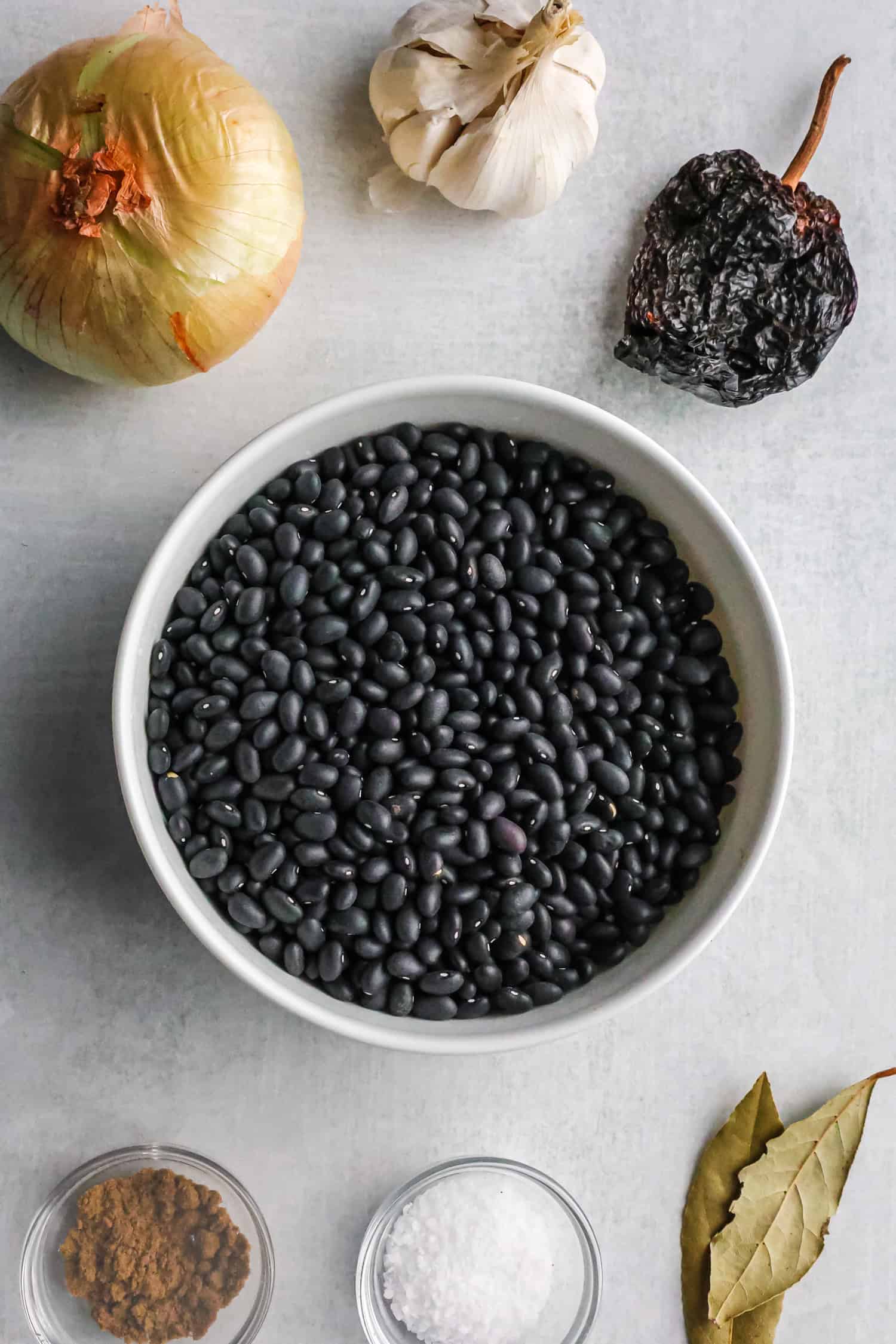 A top-down view of a white bowl filled with dried black beans surrounded by an onion, garlic, a dried chili, bay leaves, and spices.