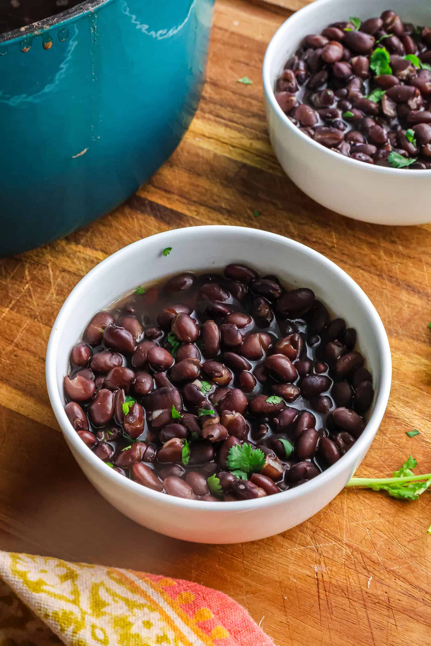 Two white bowls filled with cooked black beans garnished with fresh cilantro sitting next to a teal cooking pot on a wooden board.