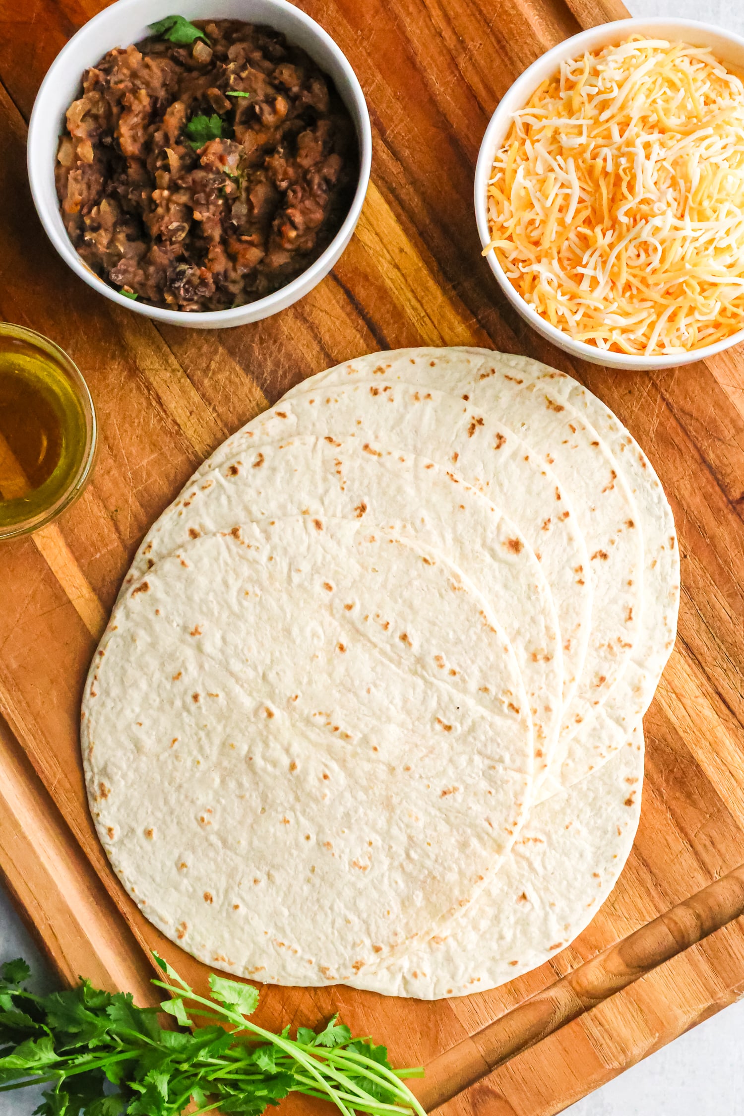 A wooden board holding a stack of flour tortillas, a bowl of refried beans, shredded cheese, and cooking oil.