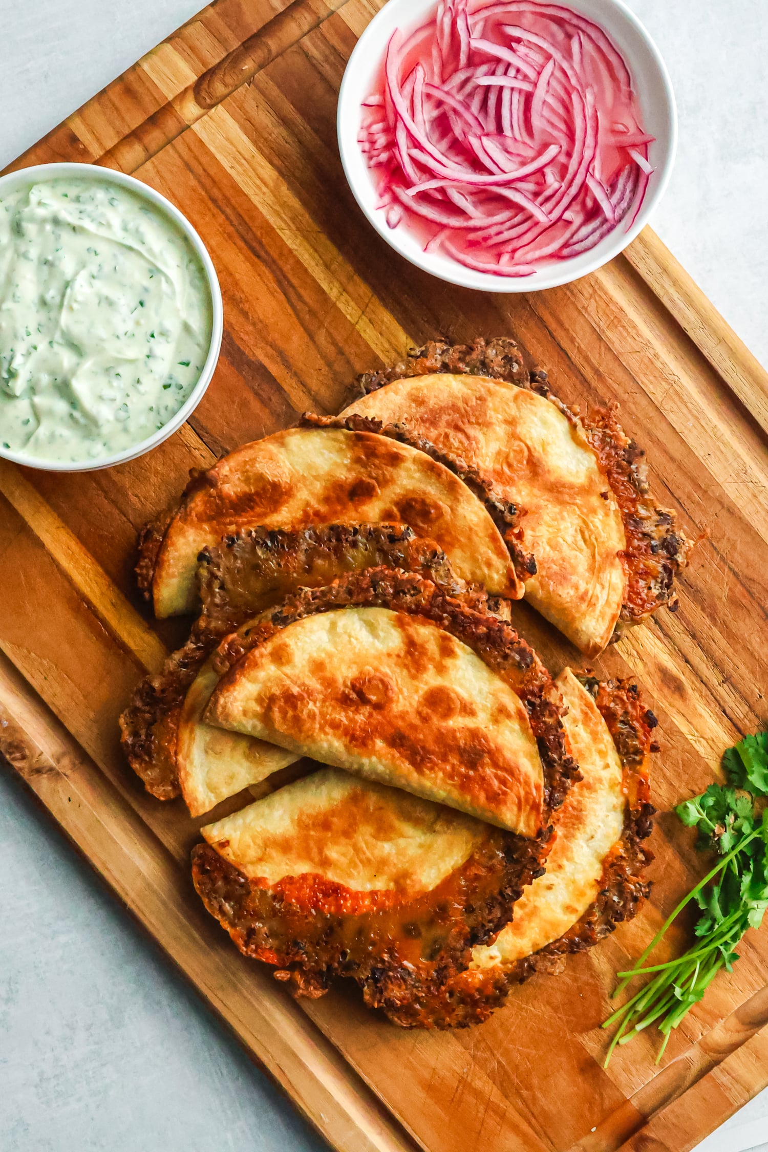 Five crispy baked tacos are served on a wooden board with bowls of green sauce and pickled red onions.