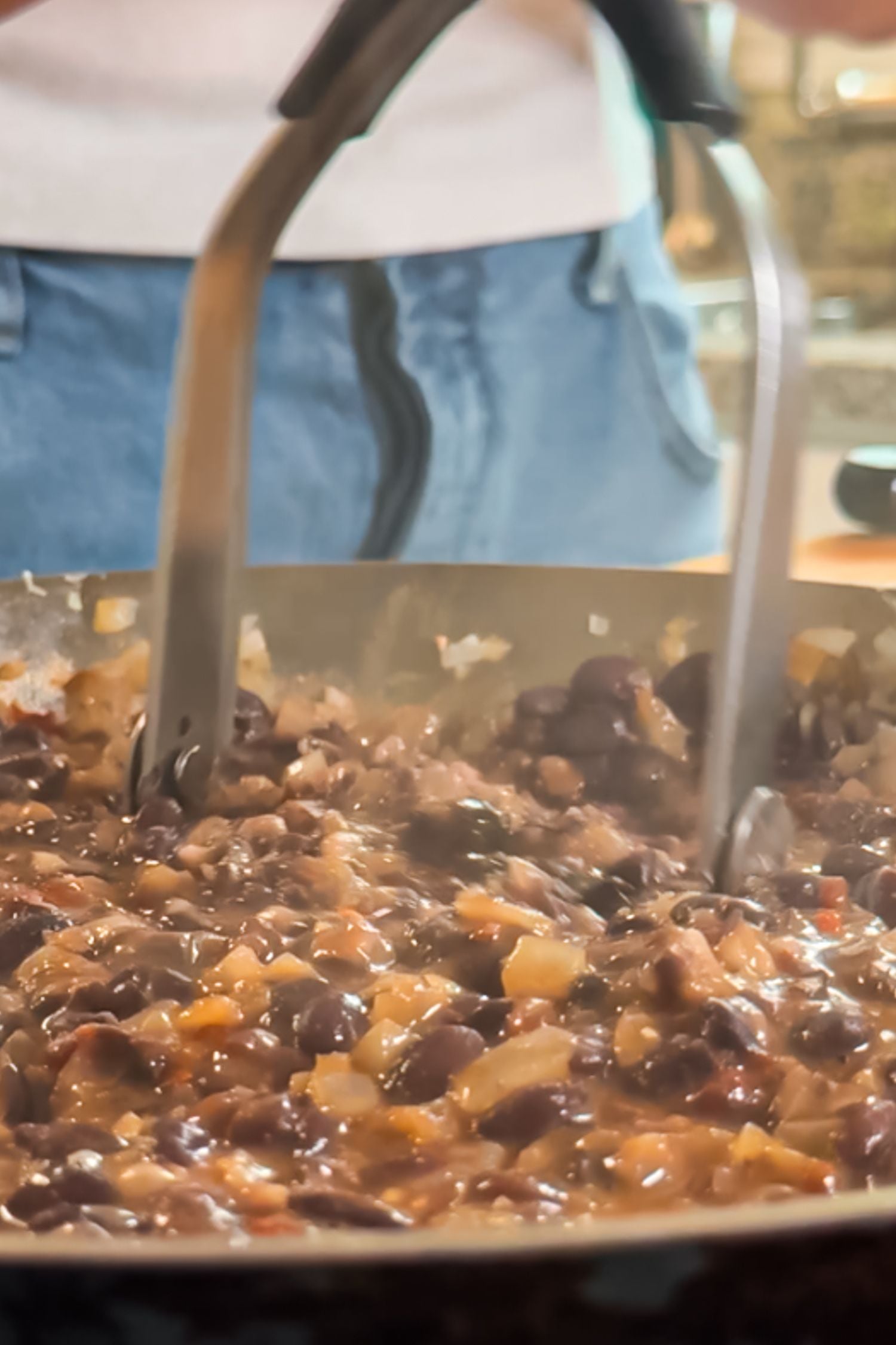 A metal potato masher is used to crush black beans and onions cooking in a pan.