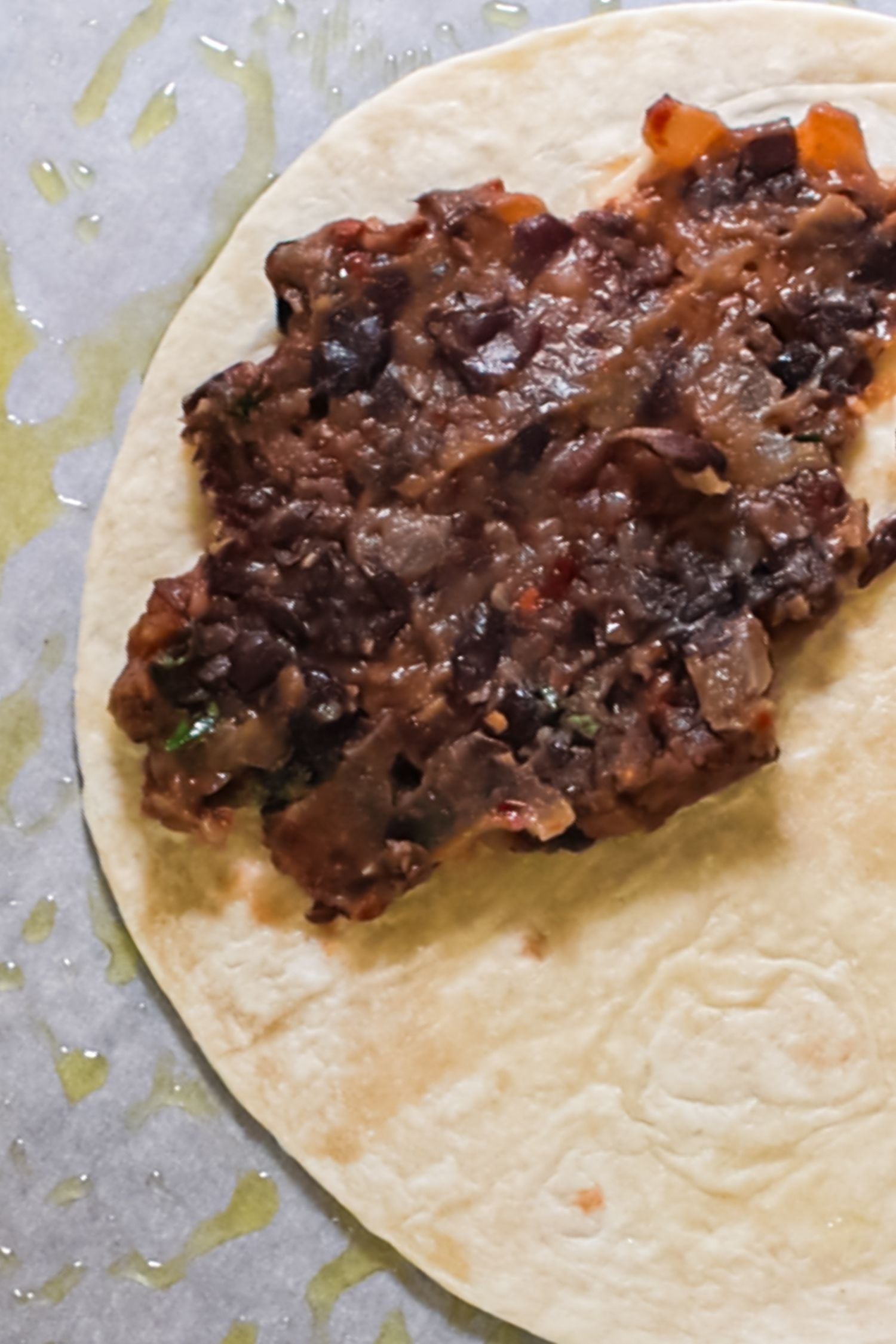 A white flour tortilla is spread with a layer of dark bean filling on a piece of parchment paper.