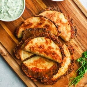 A wooden cutting board topped with crispy, golden-brown baked tacos served next to a small bowl of creamy cilantro dipping sauce.