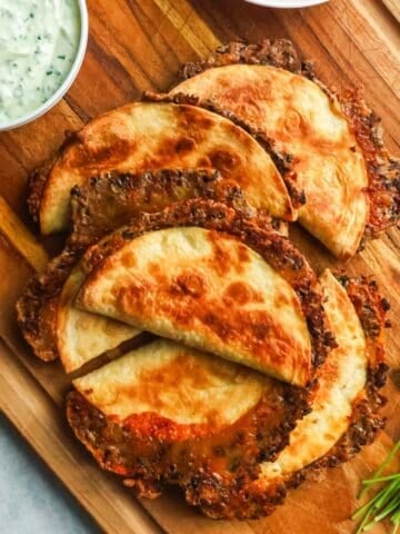 A wooden cutting board topped with crispy, golden-brown baked tacos served next to a small bowl of creamy cilantro dipping sauce.