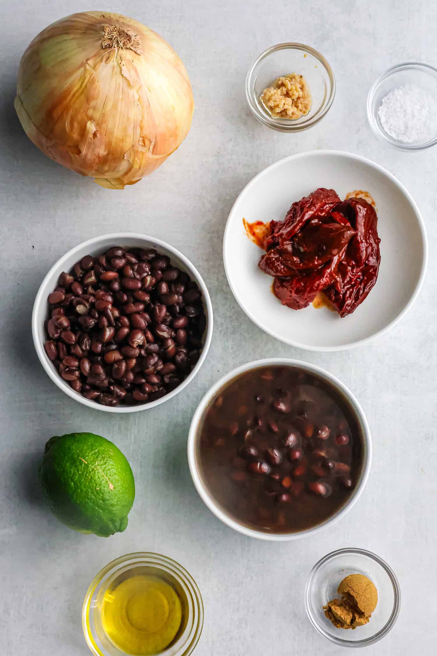 Various cooking ingredients including black beans, an onion, a lime, and spices are arranged on a grey surface.