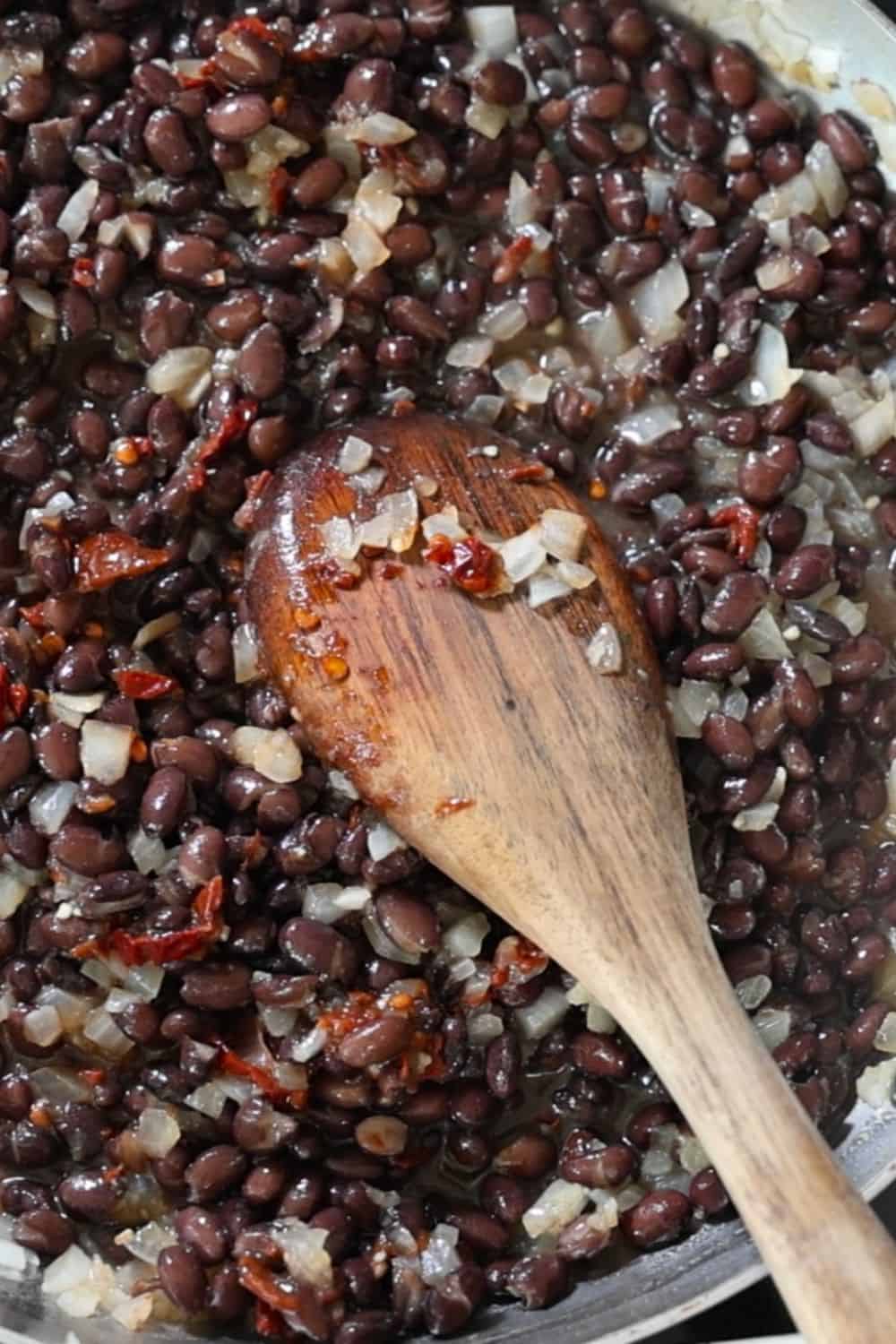 A wooden spoon stirs a mixture of black beans, diced onions, and red peppers in a cooking pan.
