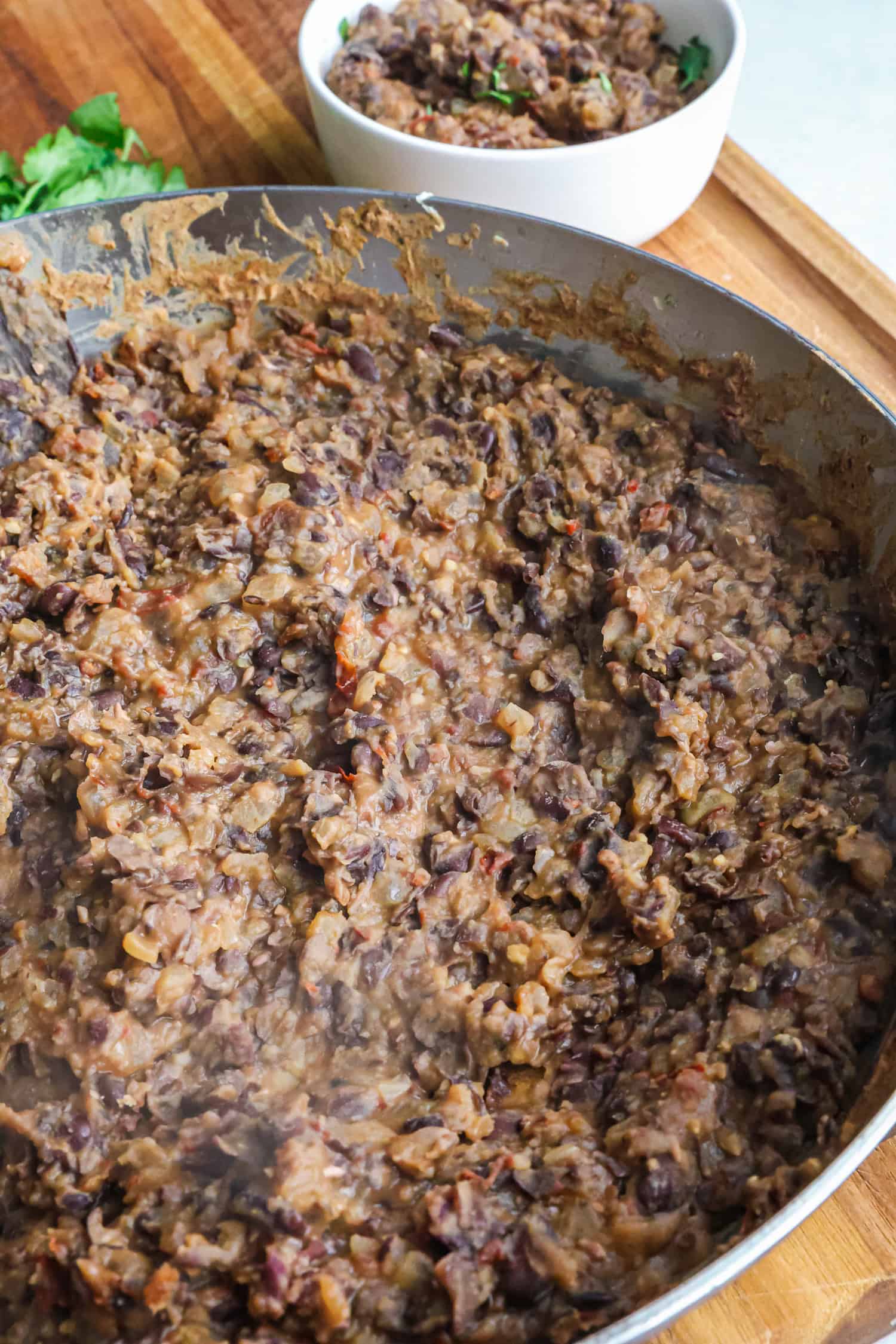 A large metal pan is filled with cooked, mashed black beans next to a small white bowl.