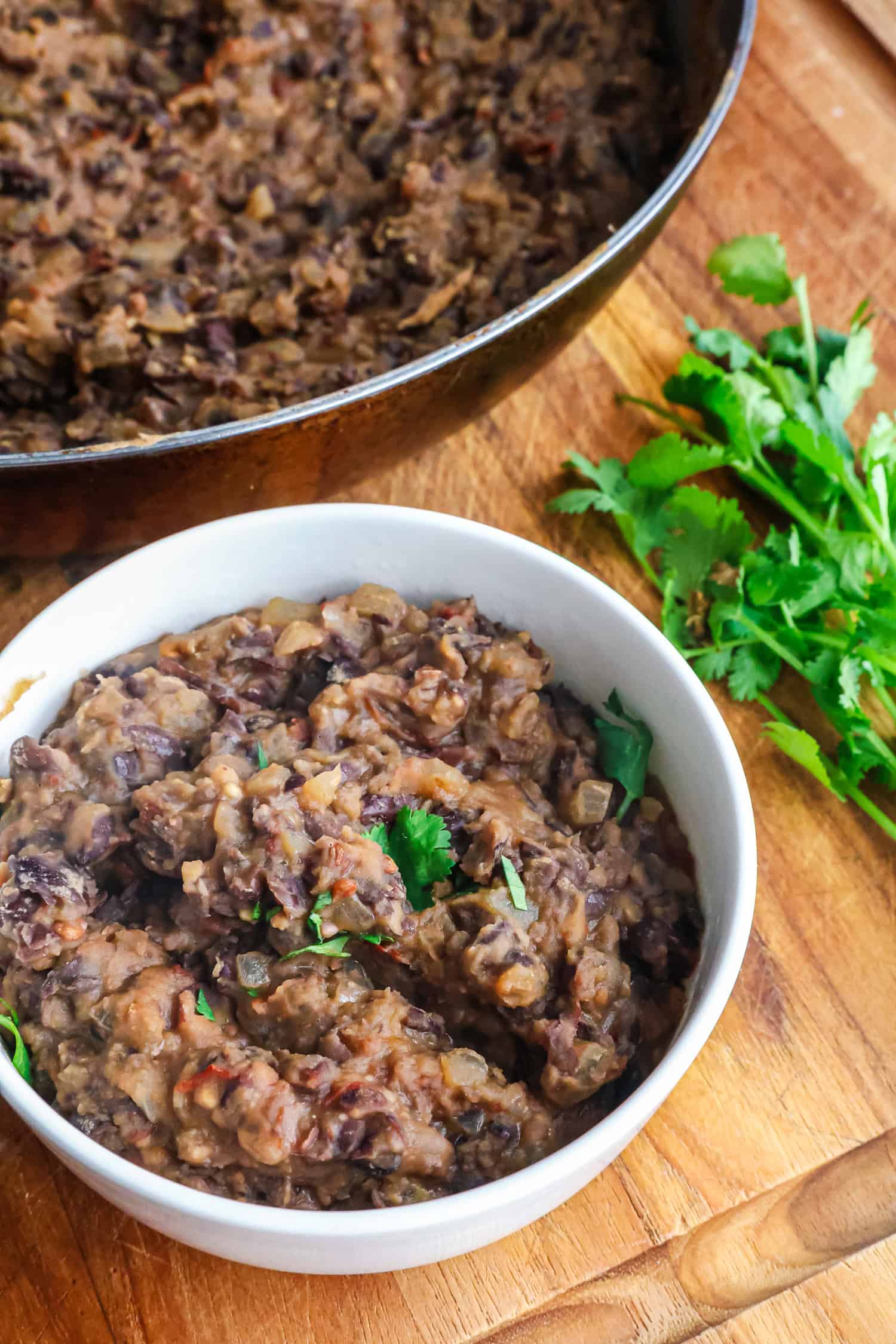 A large metal pan is filled with cooked, mashed black beans next to a small white bowl.