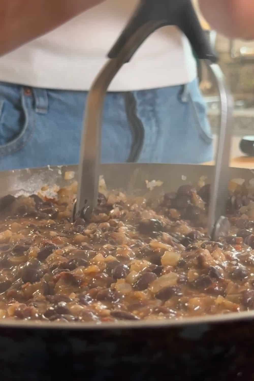 A metal potato masher is used to crush black beans and onions in a hot pan.