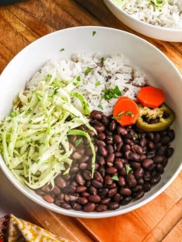 A white bowl filled with cooked black beans, white rice, shredded cabbage slaw, and sliced carrots on a wooden cutting board.