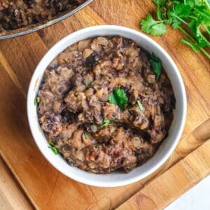 A white bowl filled with thick, mashed black beans topped with fresh cilantro on a wooden board.