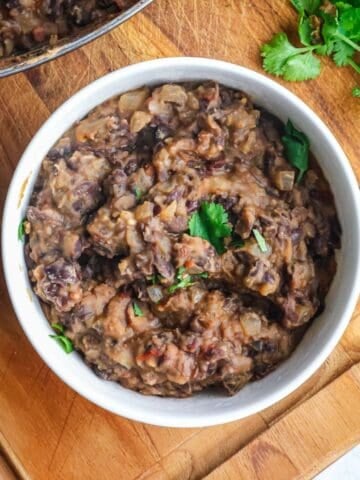 A white bowl filled with thick, mashed black beans topped with fresh cilantro on a wooden board.